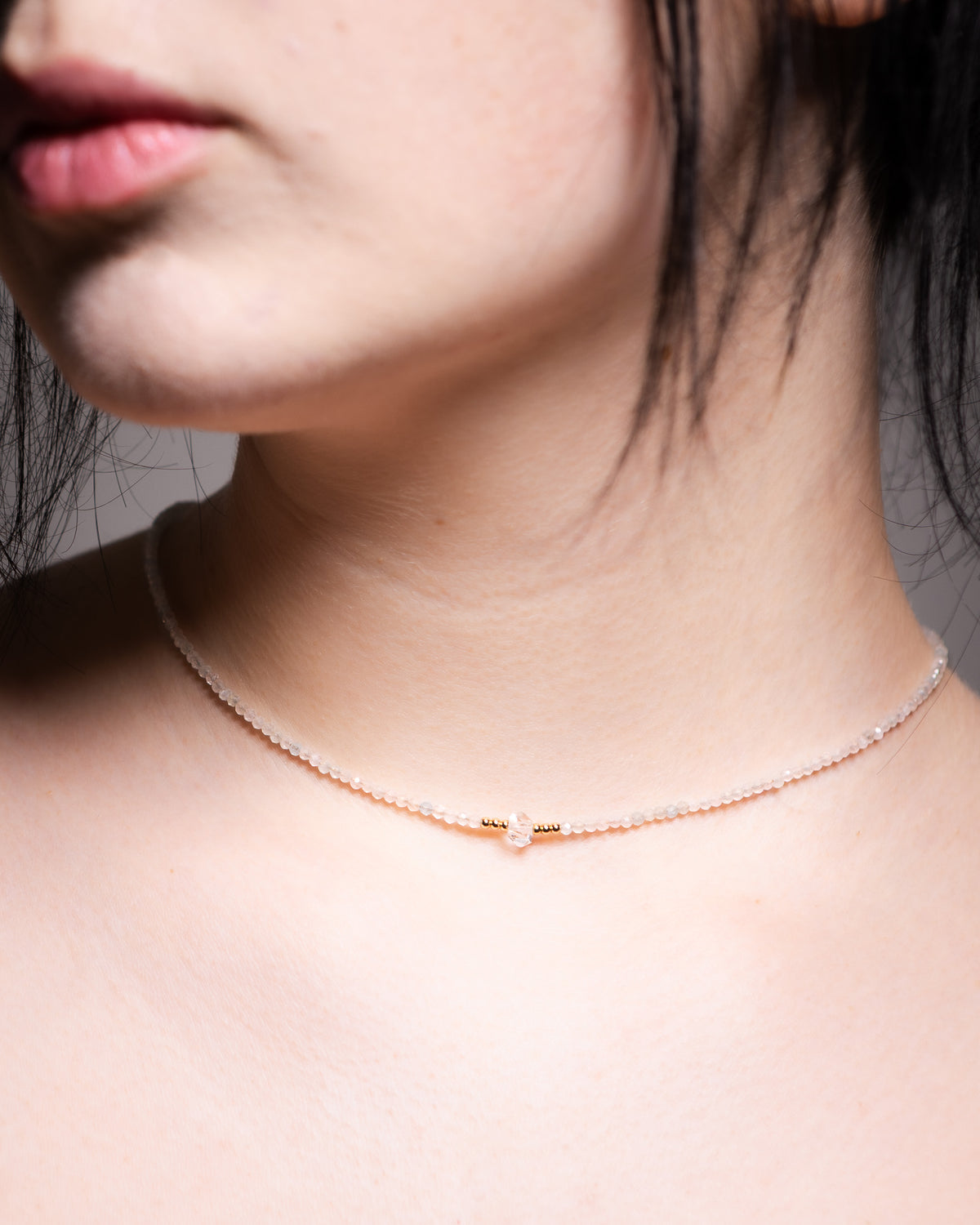 A delicate necklace worn on a model's neck, featuring a fine chain with a small gold heart charm and gold spacer beads, set against a soft gray background.